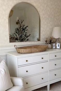 a white dresser with a basket on top of it next to a mirror and lamp