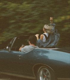 a man and woman riding in the back of a blue convertible car with trees in the background