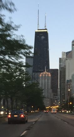 cars driving down the street in front of tall buildings at dusk with skyscrapers behind them