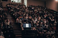 an auditorium full of people sitting and standing in front of a laptop computer on top of a table