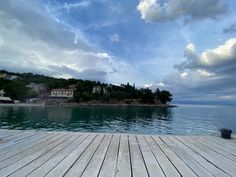 an empty dock on the water with houses in the distance and cloudy skies above it