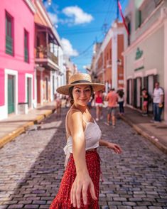 a woman in a straw hat is standing on a cobblestone street and smiling