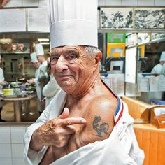 an old man in a chef's hat pointing at the camera while standing in front of a kitchen