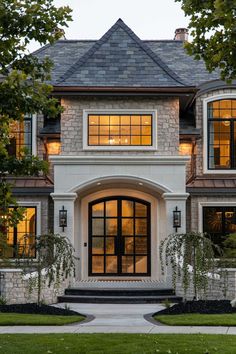 a large house with lots of windows and lights on it's front door, surrounded by greenery