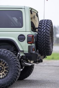 the front end of a light green jeep with black wheels and large tires, parked in a parking lot