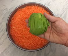 a hand holding an avocado over shredded carrots in a bowl on a table