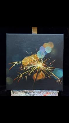 a painting with fireworks in it on top of a black surface and some lights behind it