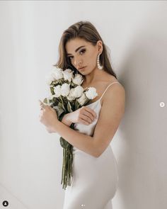 a woman in a white dress holding flowers and looking at the camera while leaning against a wall