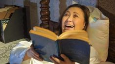 a woman laying in bed and reading a book with her mouth open while holding an open book