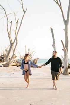 a pregnant woman holding the hand of her husband while walking on the beach with bare trees in the background