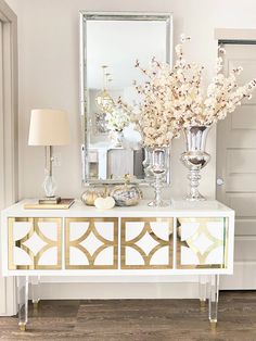 a white and gold sideboard with flowers on it in front of a large mirror