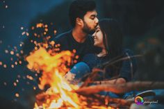 a man and woman sitting in front of a fire with their eyes close to each other