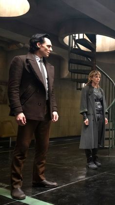 two people are standing in front of a spiral staircase and one person is wearing a suit