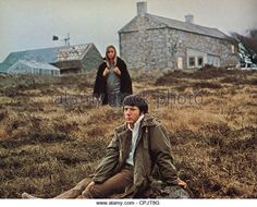 two people sitting on the ground in front of an old stone house and one person standing