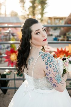 a woman in a white dress with tattoos on her arms and shoulder, holding flowers