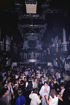 a large group of people standing around each other in a room with lights on the ceiling