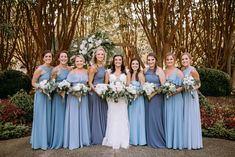 a group of women standing next to each other in front of trees and bushes holding bouquets