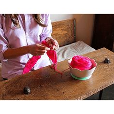 a woman crocheting pink yarn on top of a wooden table next to a bowl