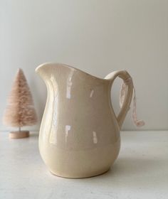 a white ceramic pitcher sitting on top of a counter next to a small tree in the corner