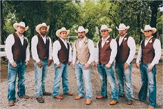 a group of men standing next to each other wearing cowboy hats and vests in front of trees