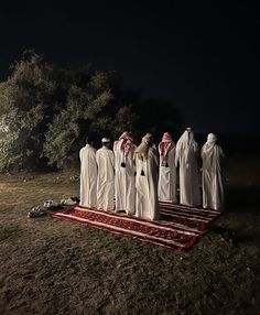 five men dressed in white robes stand on a red and black rug outside at night