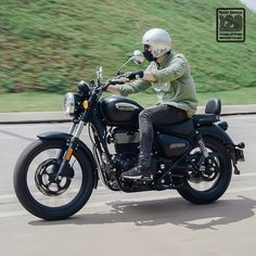 a man riding on the back of a black motorcycle down a street next to a lush green hillside