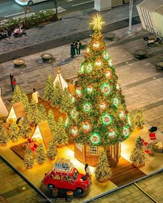 a lighted christmas tree in the middle of a street with cars parked around it and people walking by