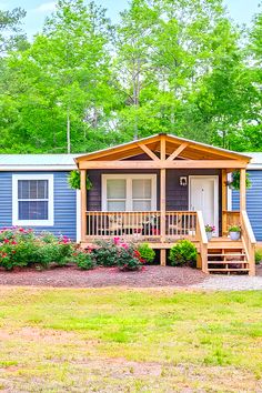 a blue mobile home in the woods with flowers and trees around it's front porch