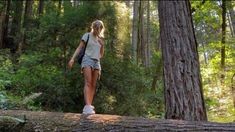 a woman walking across a fallen tree in the forest with her back to the camera