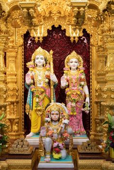 two statues of hindu god and goddess in front of a gold colored wall with flowers on it