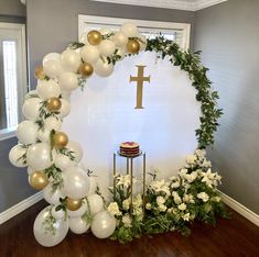 a table topped with white balloons and greenery next to a cross on the wall