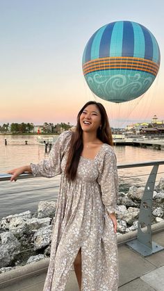 a woman in a long dress standing next to a lake with a hot air balloon above her head