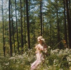 a woman standing in the middle of a forest surrounded by tall grass and trees with her hair blowing in the wind