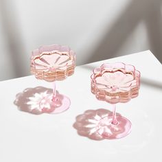 two pink wine glasses sitting next to each other on a white counter top with shadows from the glass