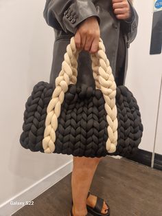 a woman holding a black and white handbag with braids on the handles, while standing in front of a wall