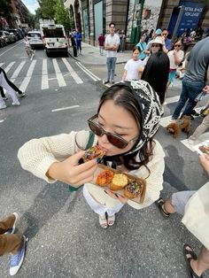 a woman eating food while standing on the side of a road with people walking by