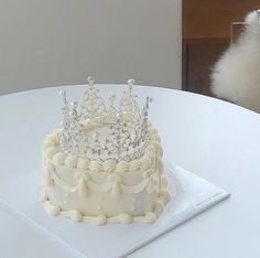 a white cake with a tiara on top sits on a table next to a cat