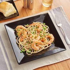 a black plate topped with pasta and shrimp on top of a wooden table next to bread