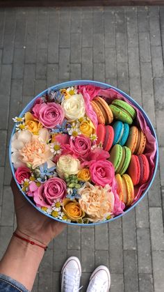 a person holding a blue bowl filled with colorful macaroons and flowers on top of a sidewalk