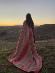 a woman wrapped up in a blanket looking out over the hills at sunset or sunrise