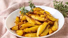 a white bowl filled with fried potatoes on top of a pink cloth next to a sprig of parsley