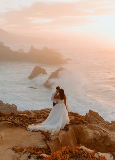 a bride and groom standing on the rocks by the ocean at sunset with waves crashing in