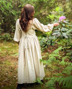 a woman in a white dress is holding out her hand and looking at a purple flower