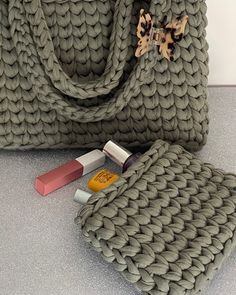 a gray knitted bag with lipstick on the bottom and two different shades of lipstick next to it