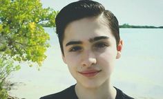 a young man standing in front of a tree and water with his eyes wide open