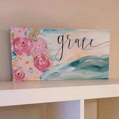 a painted canvas with the word grace on it and pink flowers sitting on a shelf