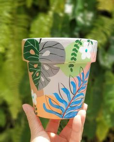 a hand holding up a cup with leaves painted on the outside and inside, in front of green foliage