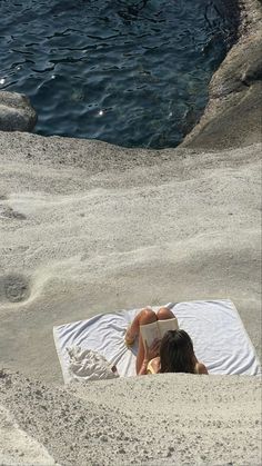 a woman laying on top of a beach next to the ocean with a book in her hand