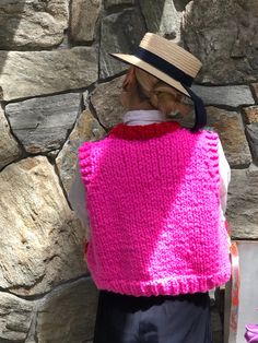 a woman wearing a pink knitted vest and hat standing in front of a stone wall