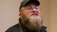 a bearded man wearing glasses and a baseball cap looks off into the distance while standing in front of a wall